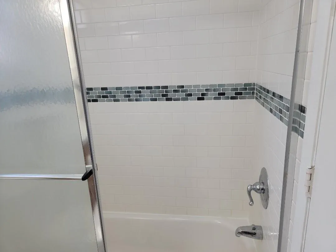 Bathtub with white subway tile surround, decorative mosaic tile accent band, polished chrome fixtures, and frosted glass shower enclosure
