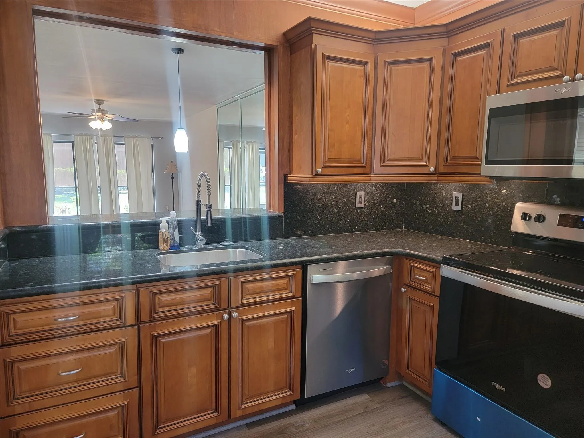 Wood-finish cabinetry with stainless steel hardware, dark countertops with matching backsplash, stainless steel appliances, and a pass-through window with a pendant light fixture