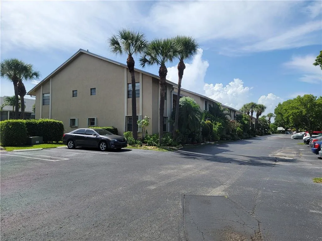 Stucco exterior with multiple windows, surrounded by mature palm trees and manicured hedges, with a paved parking area