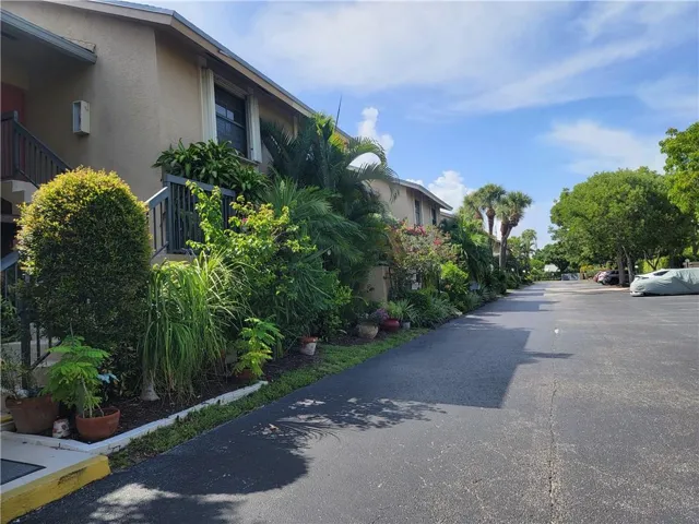 Stucco exterior building with a pitched roofline, surrounded by dense tropical landscaping, paved driveway, and mature trees