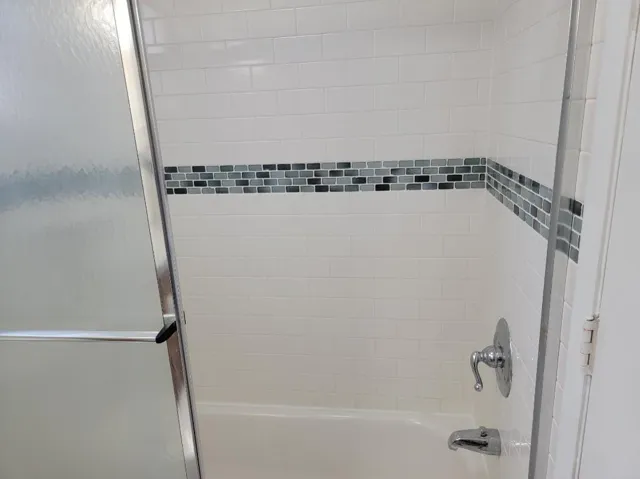 Bathtub with white subway tile surround, decorative mosaic tile accent band, polished chrome fixtures, and frosted glass shower enclosure