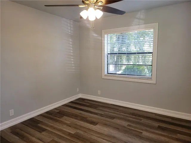 Wood-finish flooring, white trim, light gray walls, window with blinds, and a ceiling fan with integrated lighting