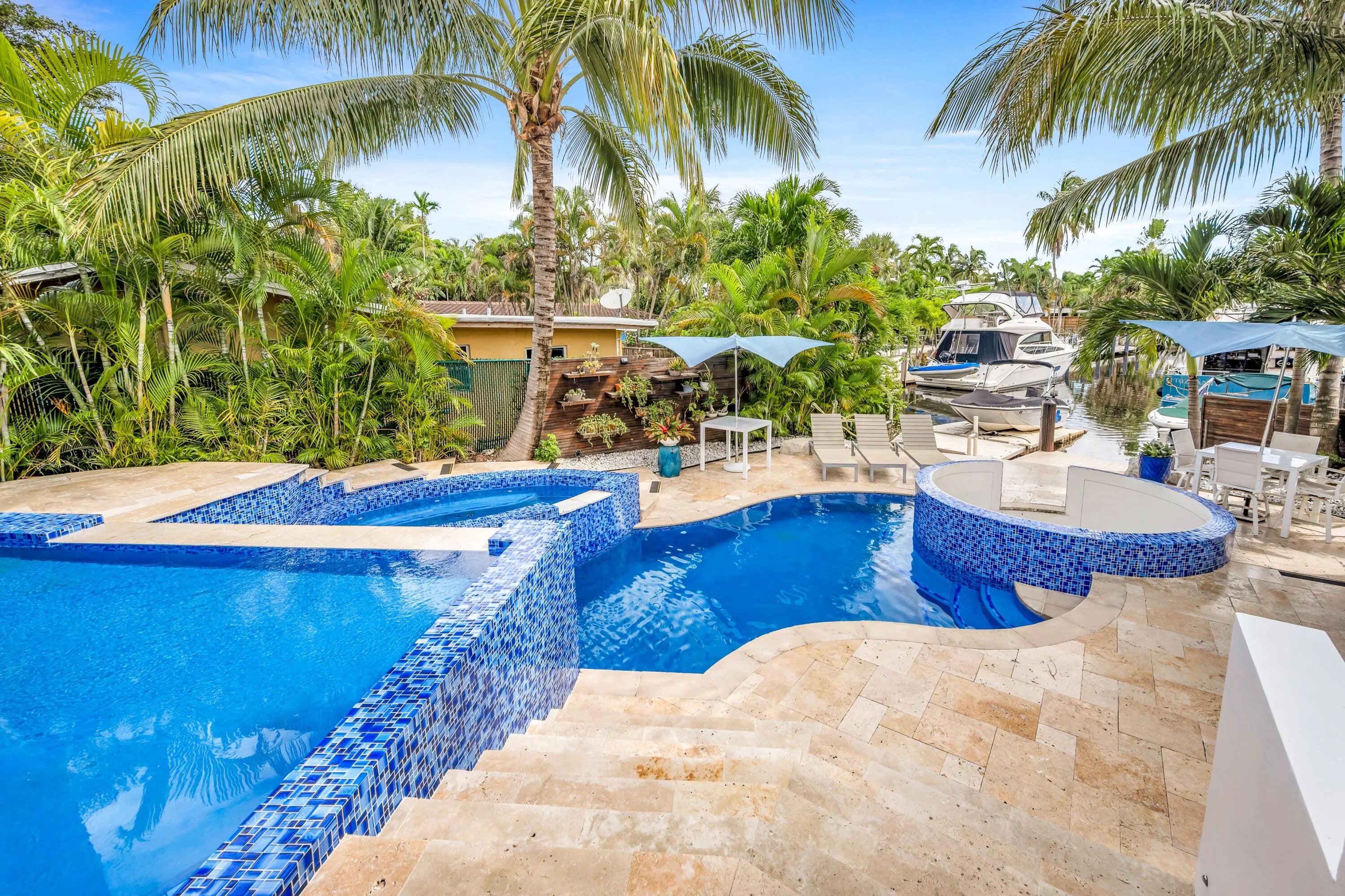 Multi-level swimming pool with blue mosaic tile accents, integrated spa, travertine paver decking, and lush tropical landscaping