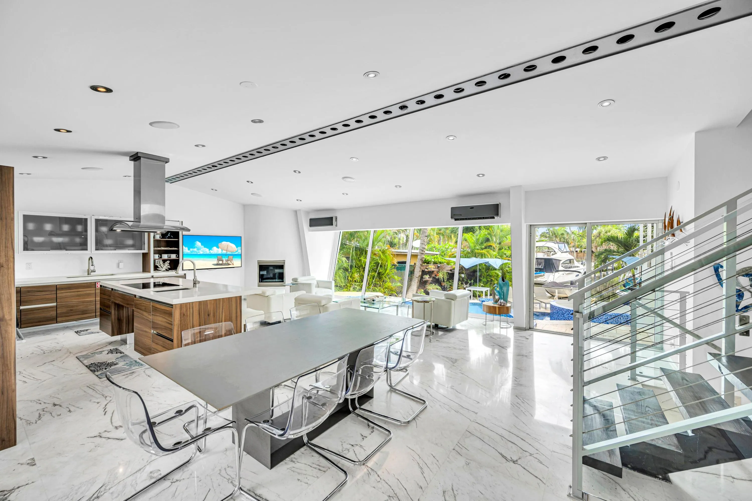 Open-concept living area with polished marble flooring, a modern kitchen island with wood-finish cabinetry, a stainless steel range hood, and a contemporary glass and metal staircase