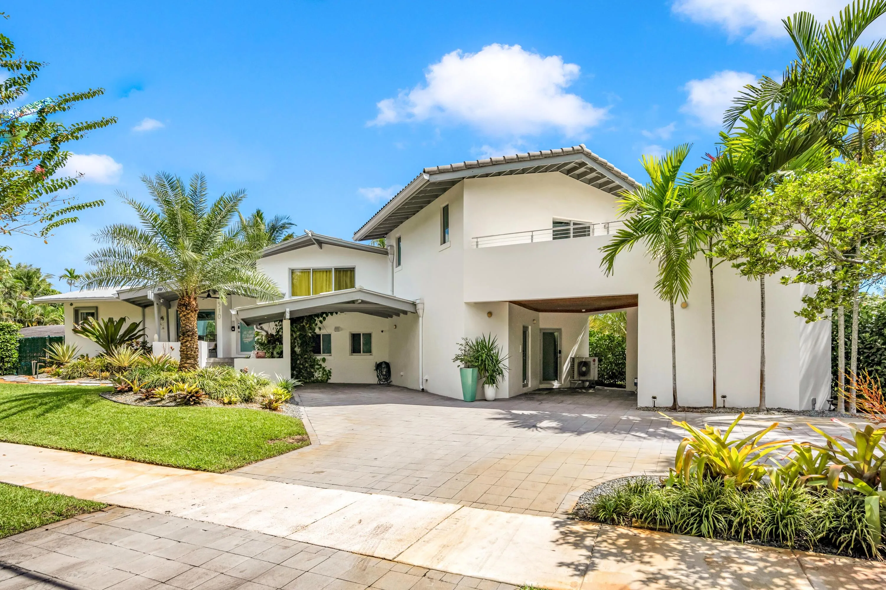 Contemporary white stucco exterior with multiple rooflines, lush tropical landscaping, and an expansive paver driveway