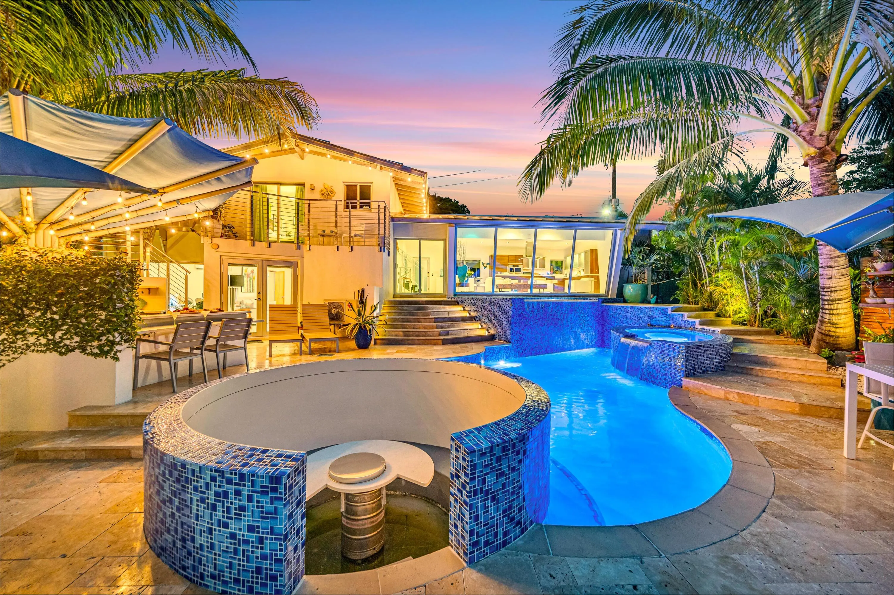 Curved swimming pool with blue tile accents, adjacent spa, integrated seating area, and multiple water features, surrounded by stone-finish paving