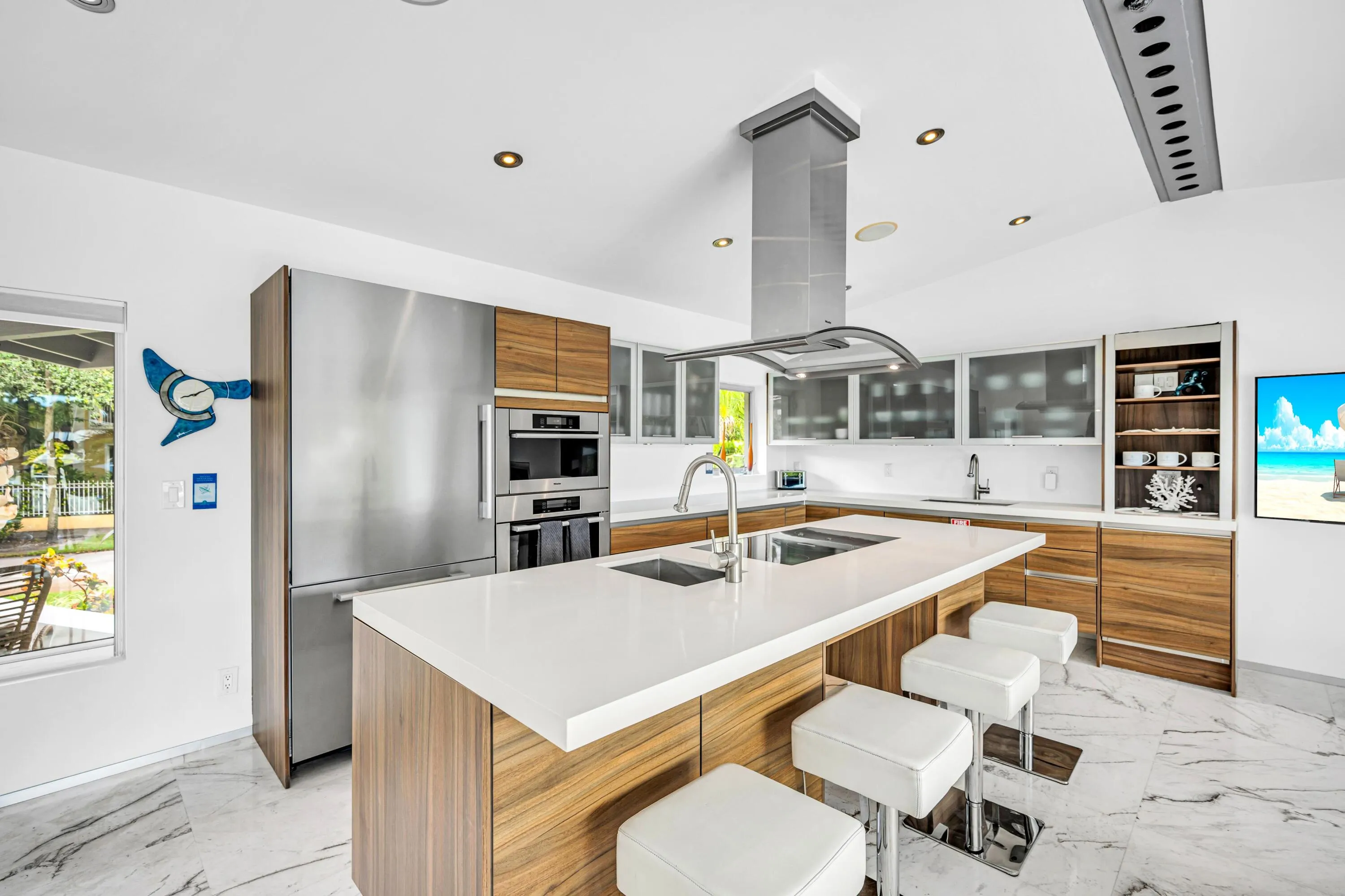 Sleek kitchen with a central island featuring dual sinks, a stainless steel range hood, built-in stainless steel appliances, wood-finish cabinetry, and white countertops