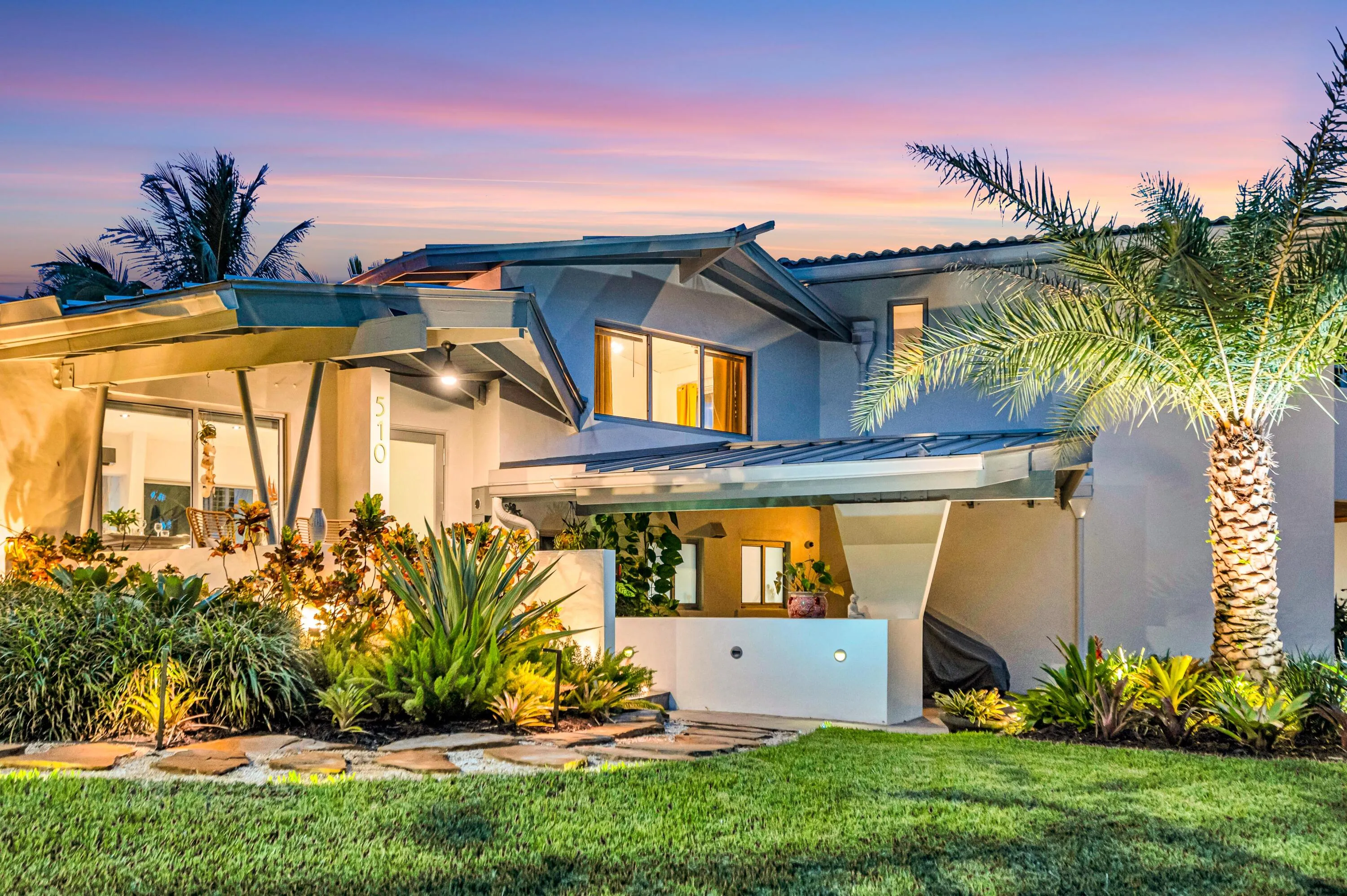 Modern exterior architecture with multiple rooflines, lush landscaping, exterior lighting, an expansive lawn, and a prominent palm tree