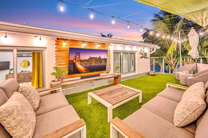 Outdoor entertainment space featuring a manicured lawn, string lighting overhead, built-in seating with wood-finish frames and cushioned upholstery, a wood-finish coffee table, and an integrated entertainment wall with a large flat-screen display and warm accent lighting