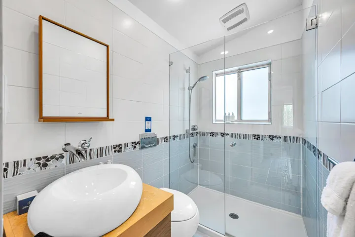 Glass-enclosed shower with a rainfall showerhead and a window, complemented by a vessel sink atop a wood-finish vanity, and a wall-mounted mirror with a wood frame
