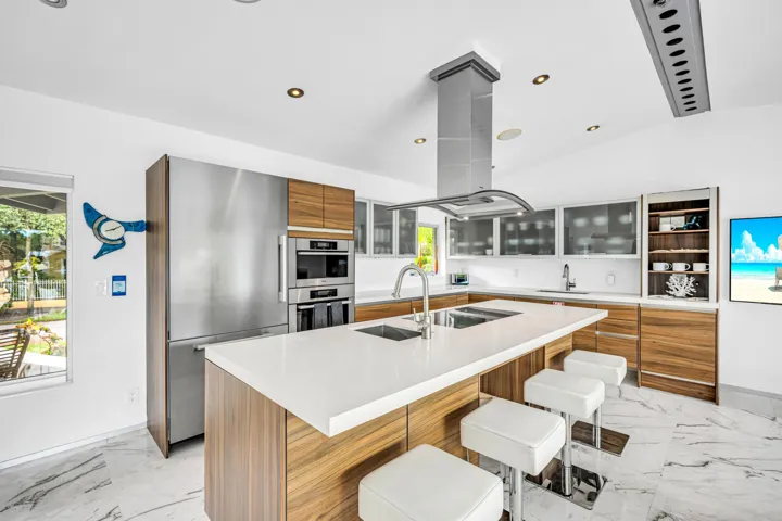 Sleek kitchen with a central island featuring dual sinks, a stainless steel range hood, built-in stainless steel appliances, wood-finish cabinetry, and white countertops