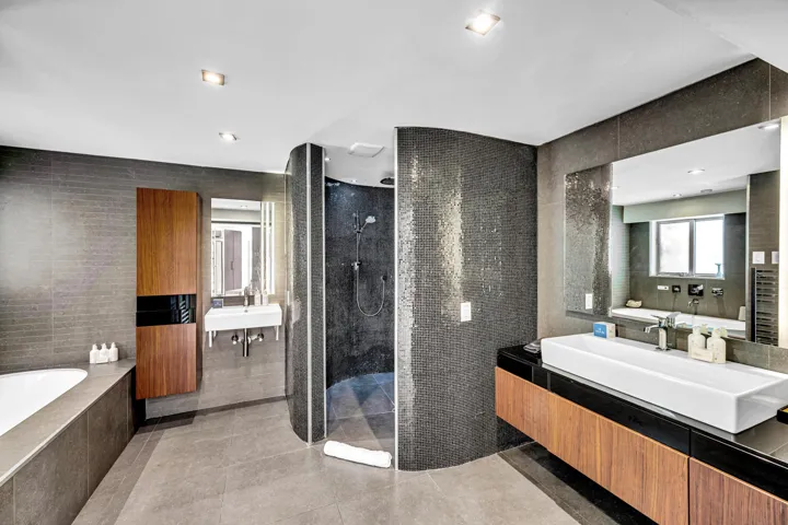 Curved tile shower enclosure with dark mosaic tile, dual wall-mounted sinks, wood-finish vanity with a vessel sink, recessed lighting, and a built-in soaking tub