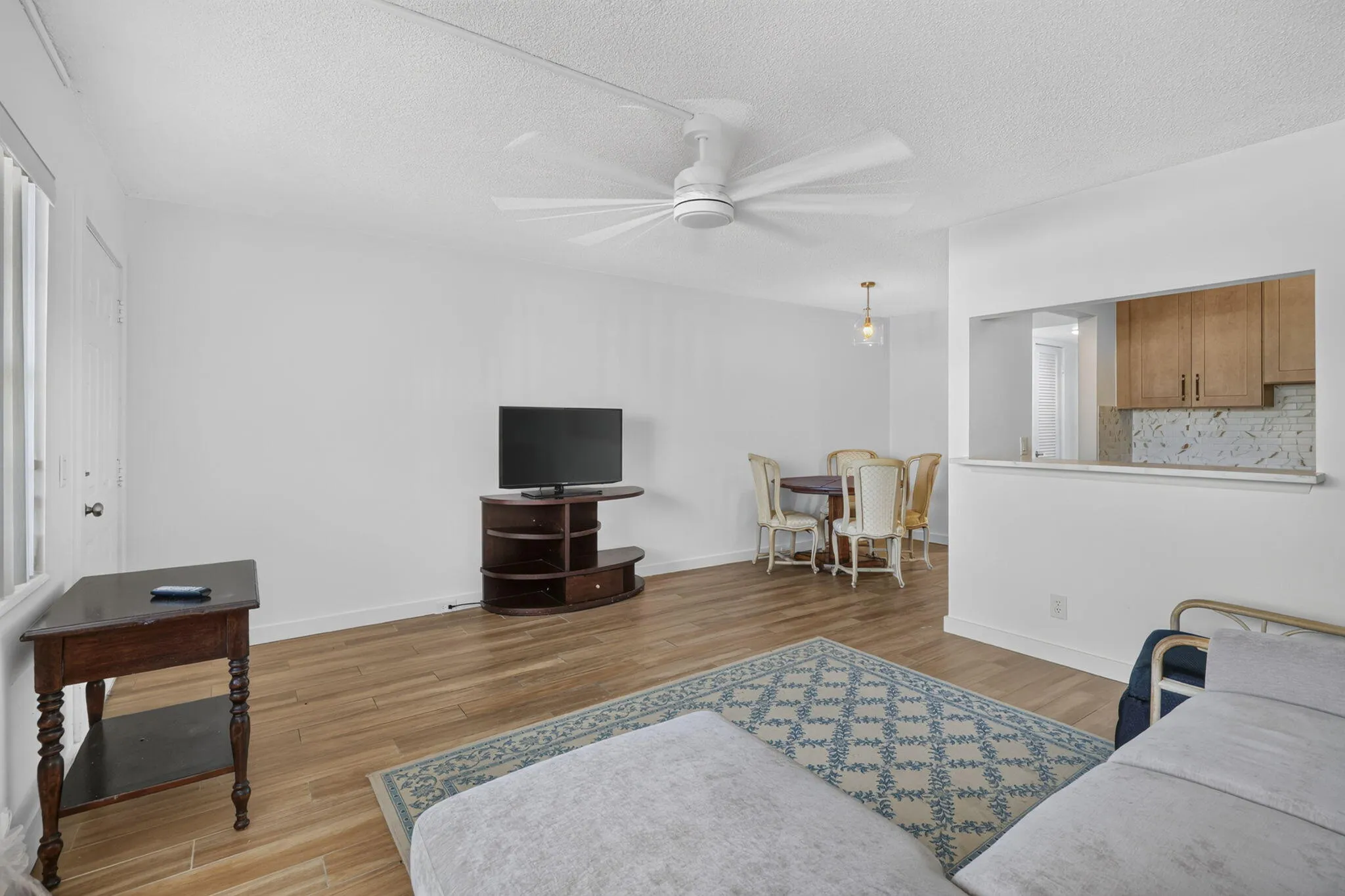 Open-concept living area featuring wood-finish flooring, a ceiling fan, and a pass-through window to the kitchen with wood cabinetry and a tiled backsplash