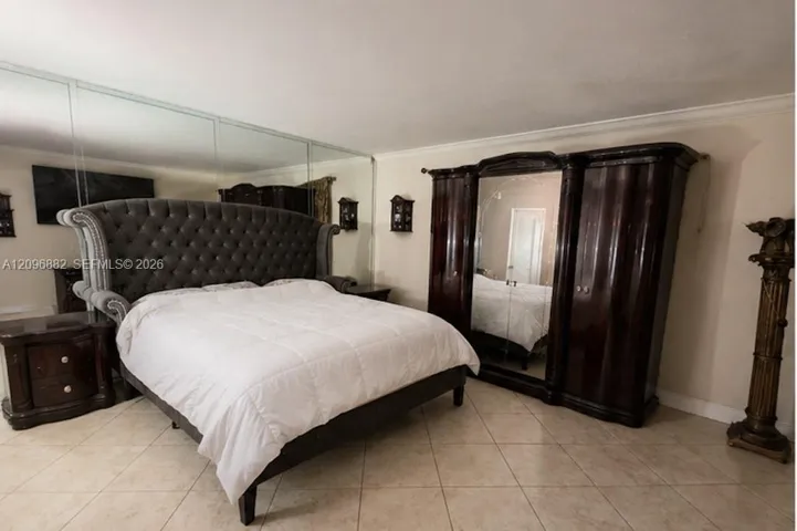 Spacious primary bedroom featuring a mirrored accent wall, tile flooring, crown molding, and ample room for substantial furnishings.