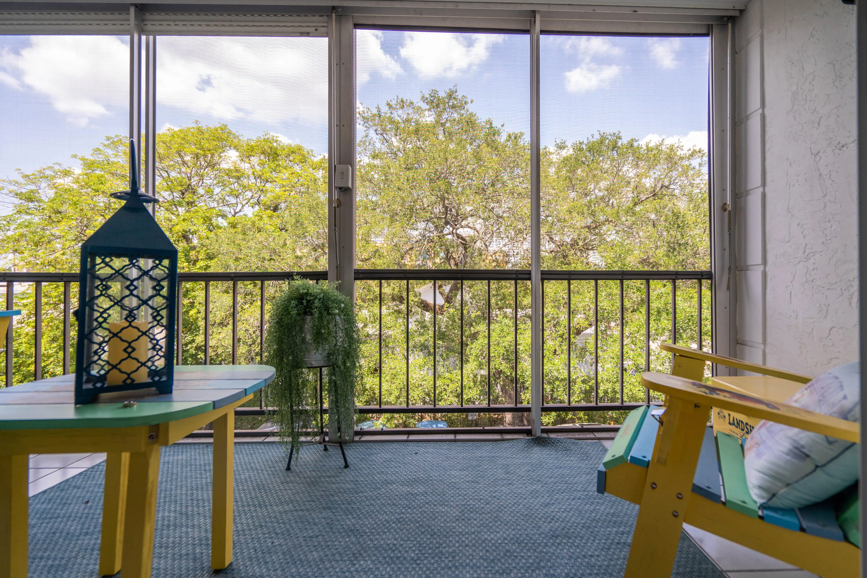 Elevated balcony with floor-to-ceiling glass doors framing lush tree canopy, featuring steel railing, wood-finish dining table, and carpet flooring