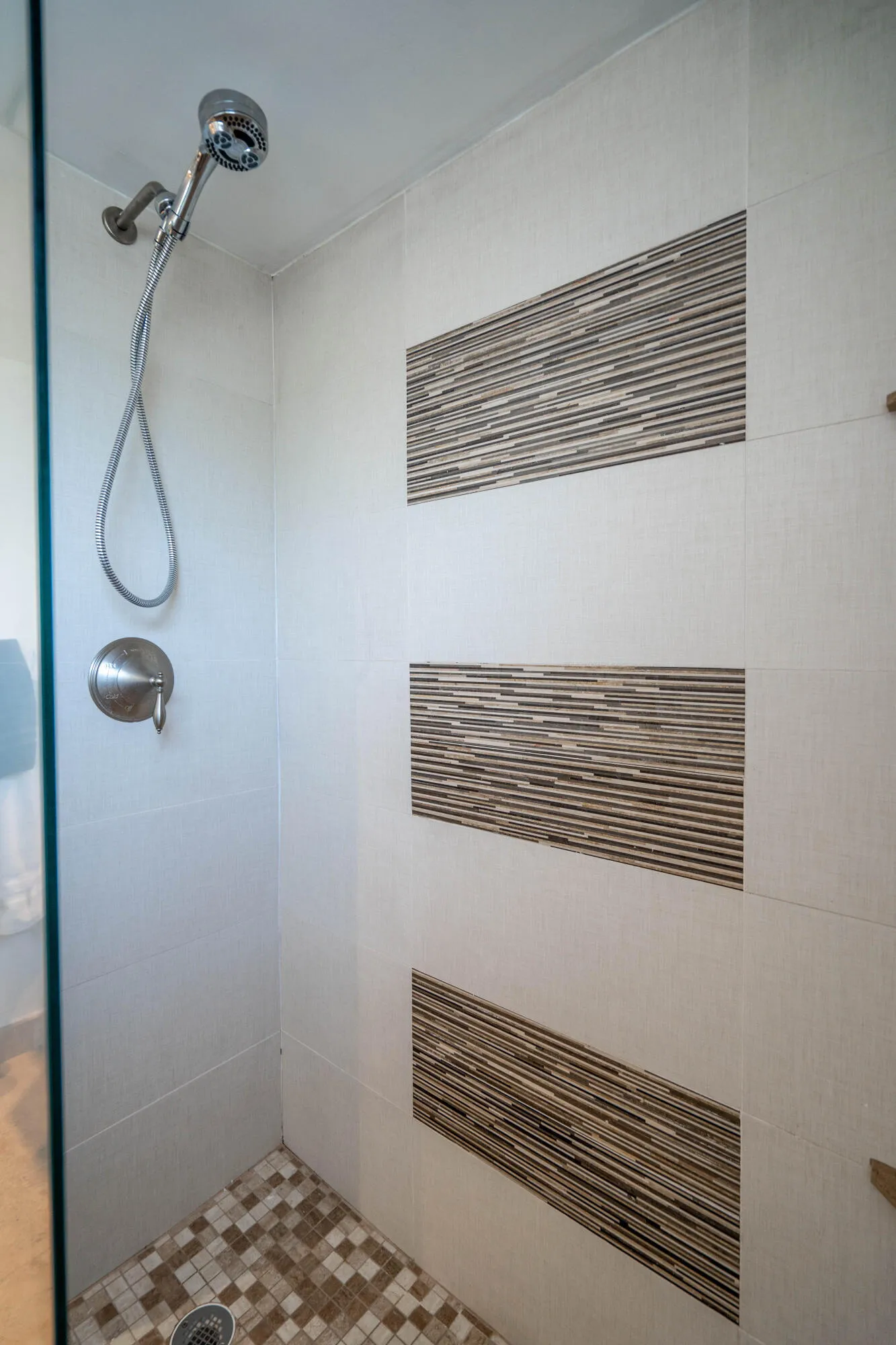 Shower with a chrome handheld showerhead, white wall tiling, decorative striped accent tiles, and mosaic floor tiling