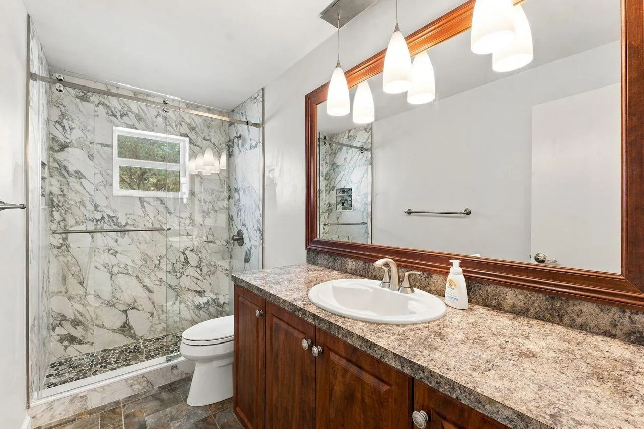 Walk-in shower with marble-style tile surround and pebbled floor, vanity with wood-finish cabinetry, stone-look countertop, and large framed mirror