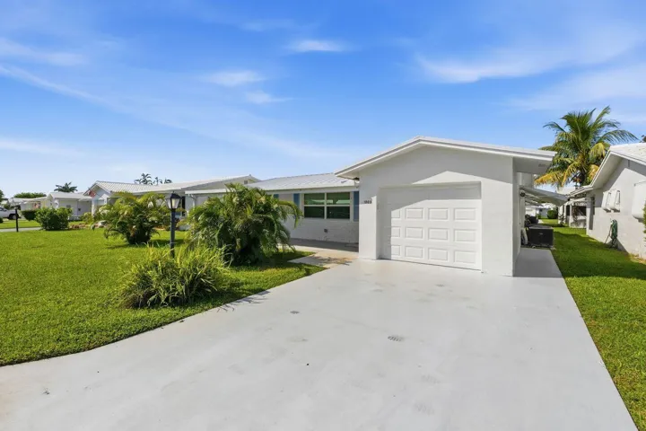 White stucco exterior, attached garage, expanded concrete driveway, manicured lawn, and mature landscaping