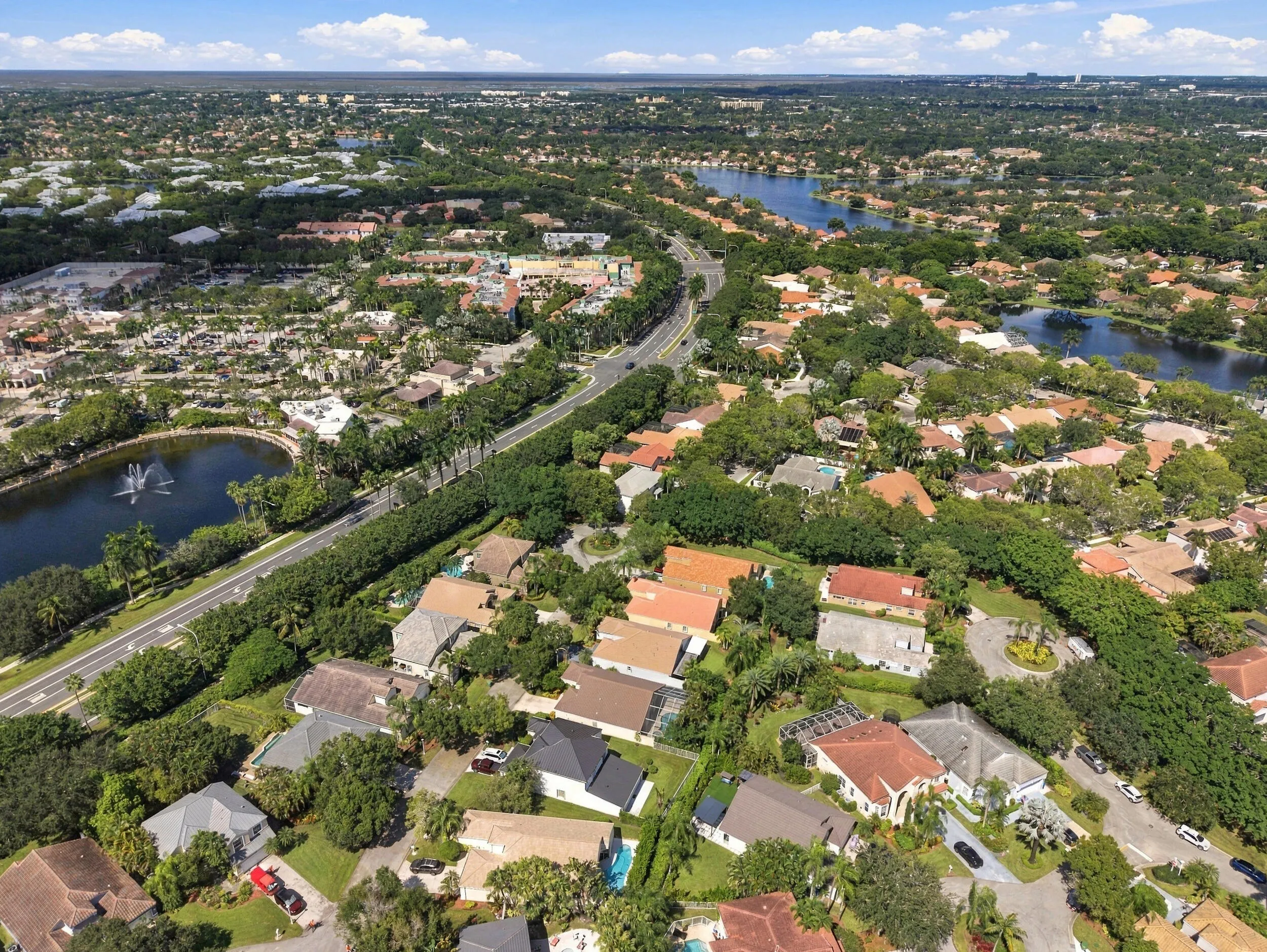 Aerial perspective showcasing a residential area with varied rooflines, abundant mature trees, multiple water features including lakes with fountains, and an extensive road network