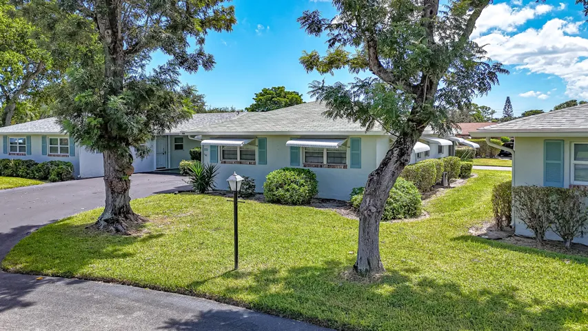 Light-toned exterior with blue shutters, white awnings, mature trees, manicured lawns, and asphalt driveways