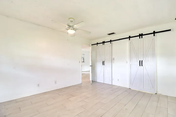 White wood-finish flooring, two sets of white barn doors with black hardware, and a ceiling fan with integrated lighting