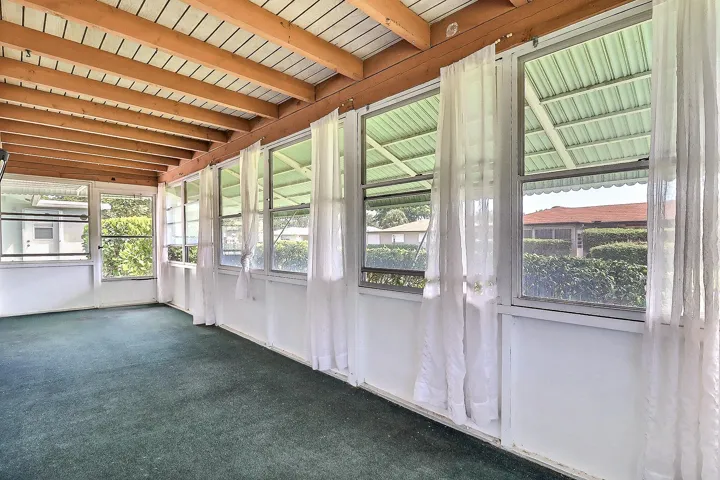 Enclosed porch with exposed wood beam ceiling, white paneled walls, multiple windows, and green carpeting