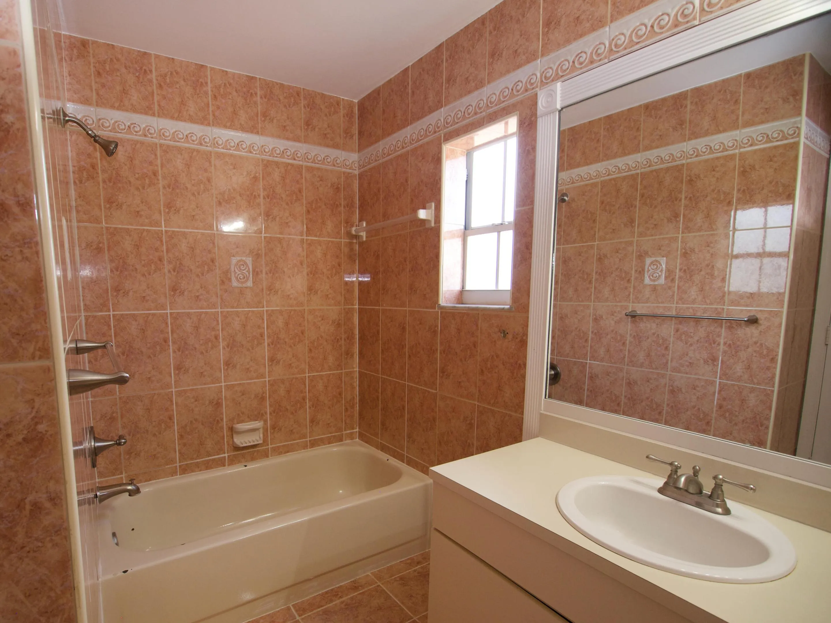 Bathroom featuring extensive tile work, a built-in bathtub with shower fixtures, a single vanity with an integrated sink, a large wall-mounted mirror, and a window