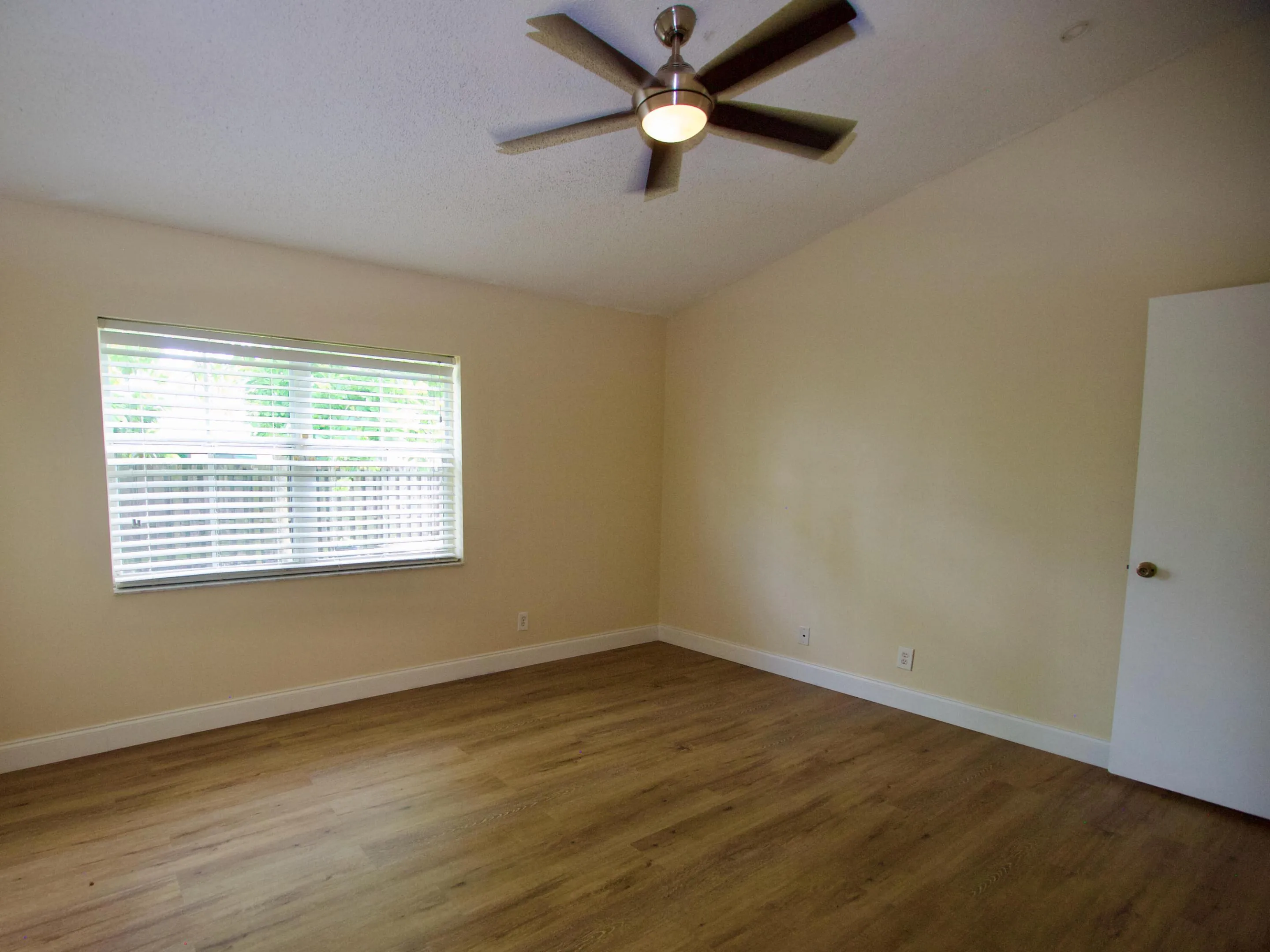Wood-finish flooring, neutral wall tones, window with horizontal blinds, and a ceiling fan with integrated lighting