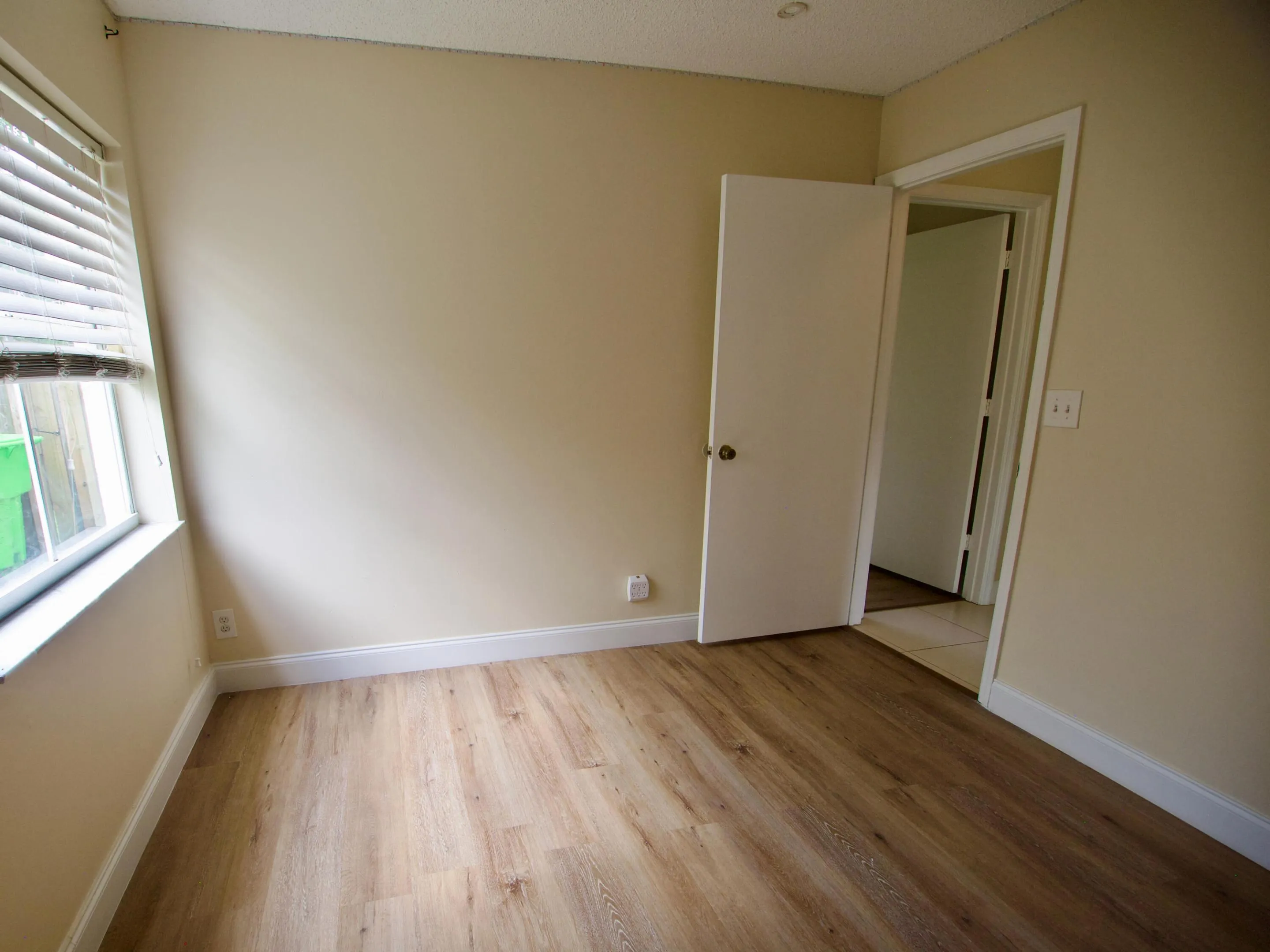 Wood-finish flooring, neutral wall tones, window with horizontal blinds, white interior door, and white trim throughout