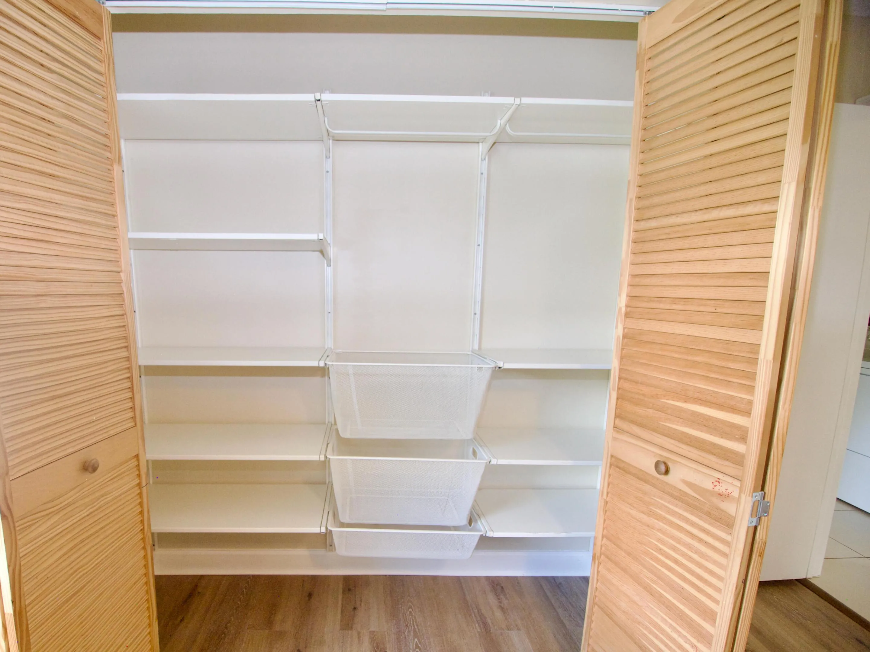 Slatted bi-fold doors, wood-finish flooring, adjustable white shelving, and pull-out storage baskets