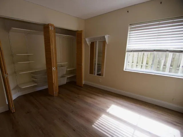 Wood-finish flooring, built-in closet organization system, louvered bi-fold closet doors, window with blinds, and a wall-mounted mirror