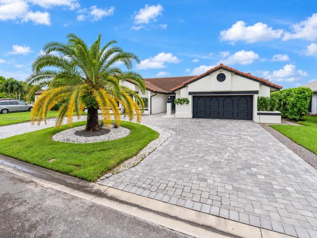 Single-story exterior featuring a light-colored stucco façade, dark-toned garage door with decorative paneling, terracotta-style roof tiles, and an expansive paved stone driveway