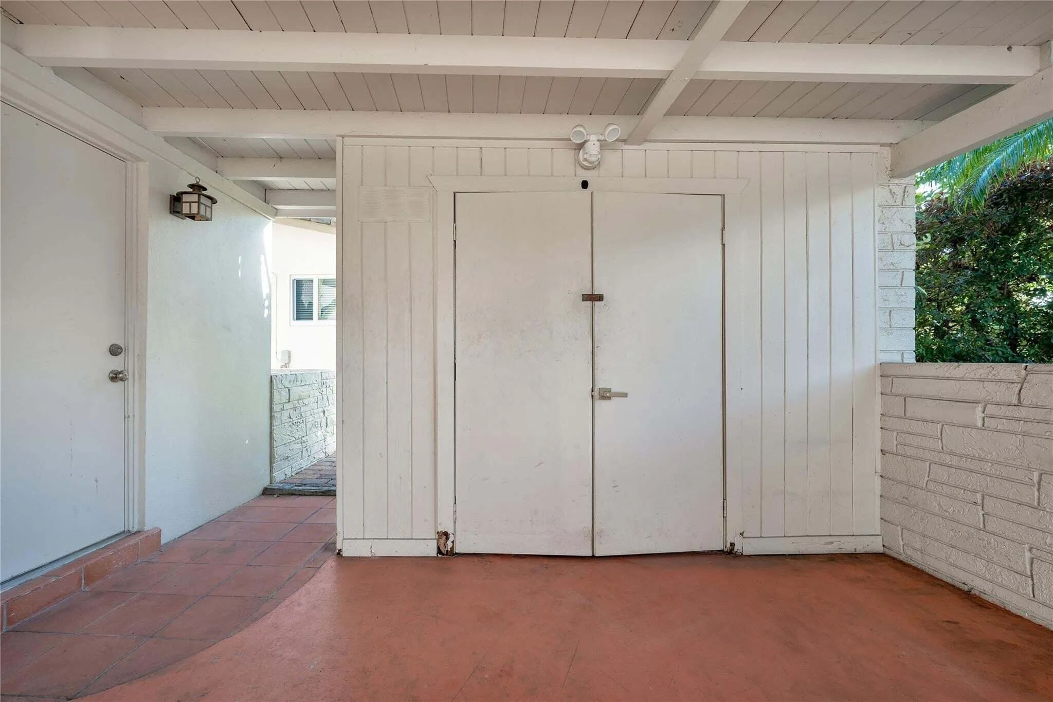 Covered outdoor area with terracotta-toned flooring, white exposed beam ceiling, and a white paneled storage unit