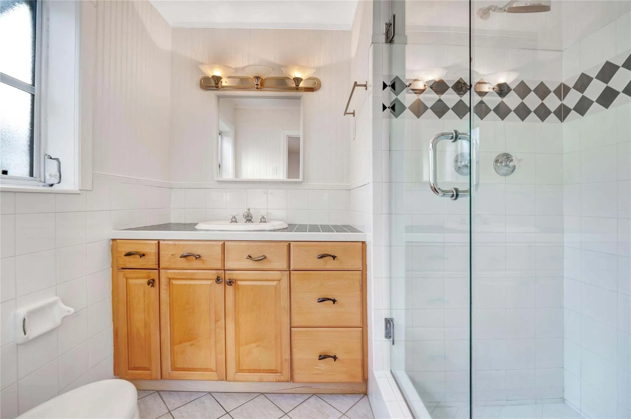 White tiled bathroom featuring a wood-finish vanity with a built-in sink, a glass-enclosed shower with a decorative tile band, and a vanity light fixture