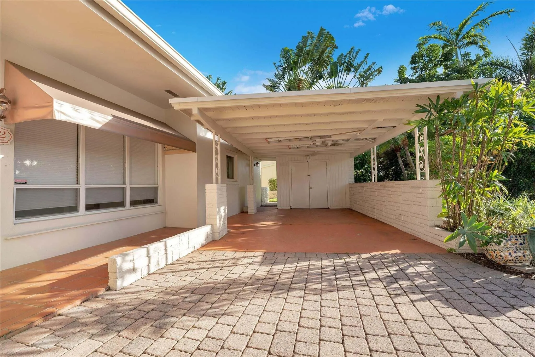 Paved brick driveway leading to a covered carport, featuring a white brick planter and lush landscaping