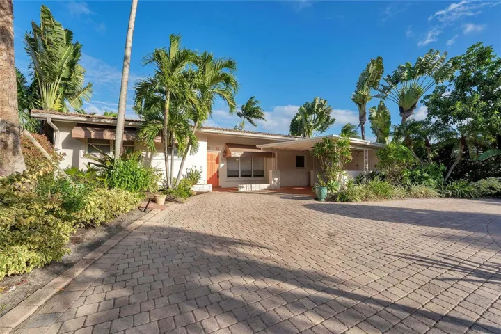Expansive paver driveway leading to a residence with a covered entryway, decorative awnings, and mature tropical landscaping