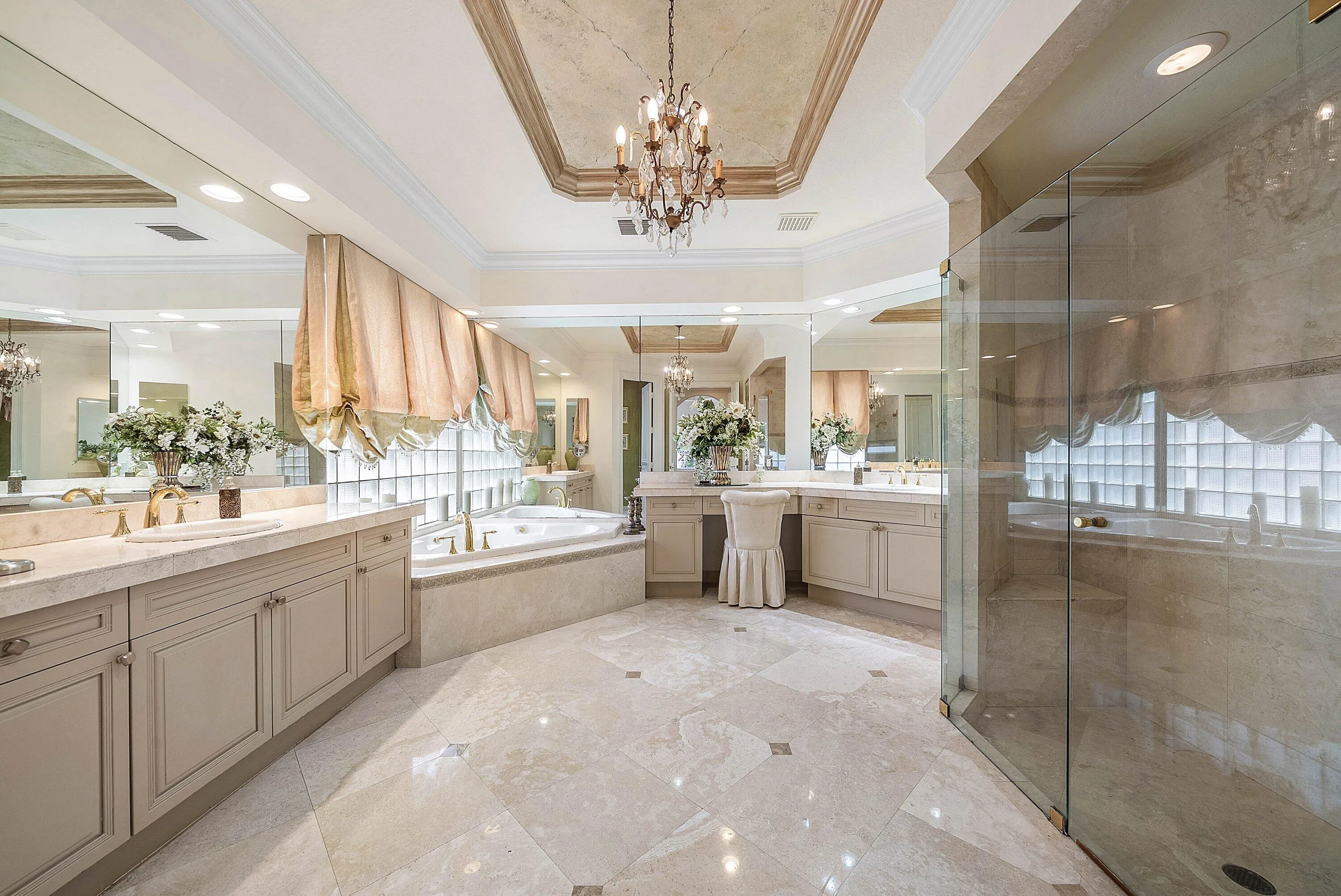 Marble flooring with accent details, dual vanities with integrated sinks, recessed jetted bathtub, glass-enclosed shower, and a decorative tray ceiling with a chandelier