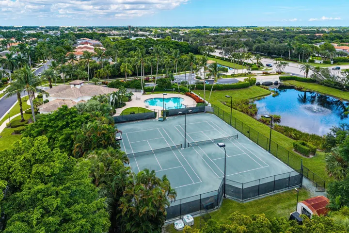 Two Resurfaced Har-Tru like tennis courts with perimeter fencing and floodlights for evening tennis, a swimming pool with a surrounding paved deck, a pond with a water feature, and extensive landscaping with mature trees