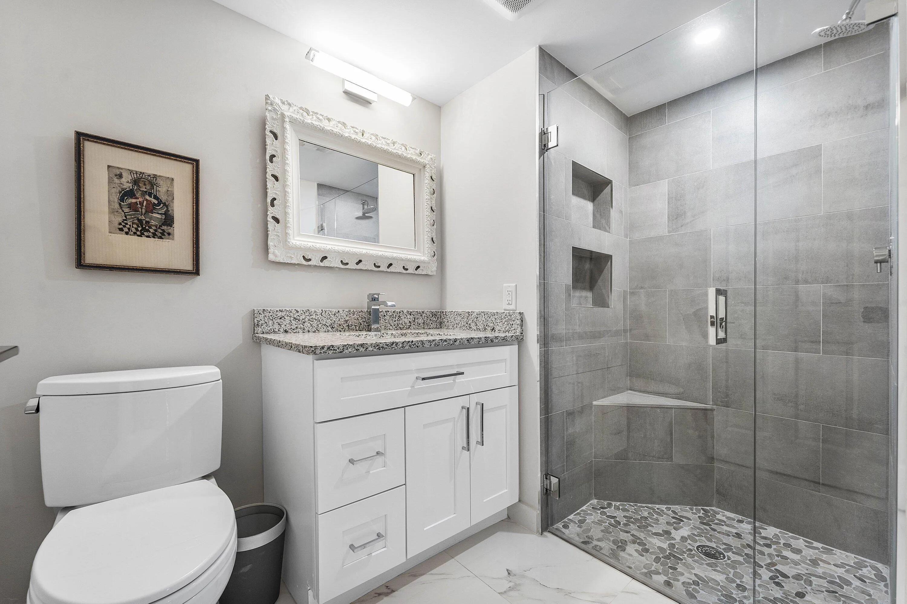 White vanity with granite countertop, glass-enclosed shower, tiled shower walls with recessed niches, pebble-tiled shower floor, and tile flooring