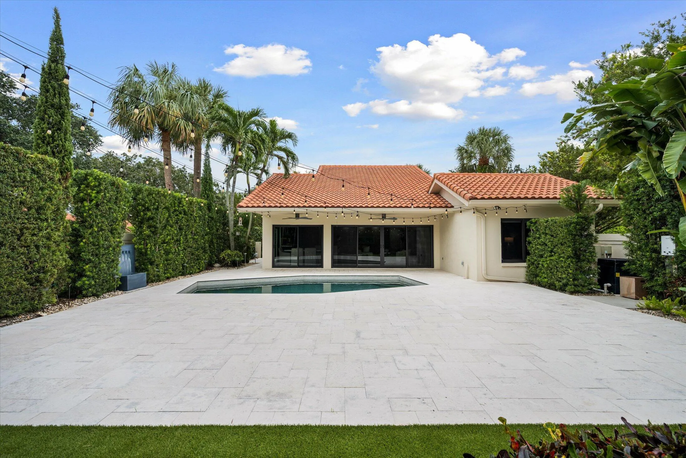 Terracotta tile roof, expansive paver patio, integrated swimming pool, mature privacy hedges, and lush tropical landscaping