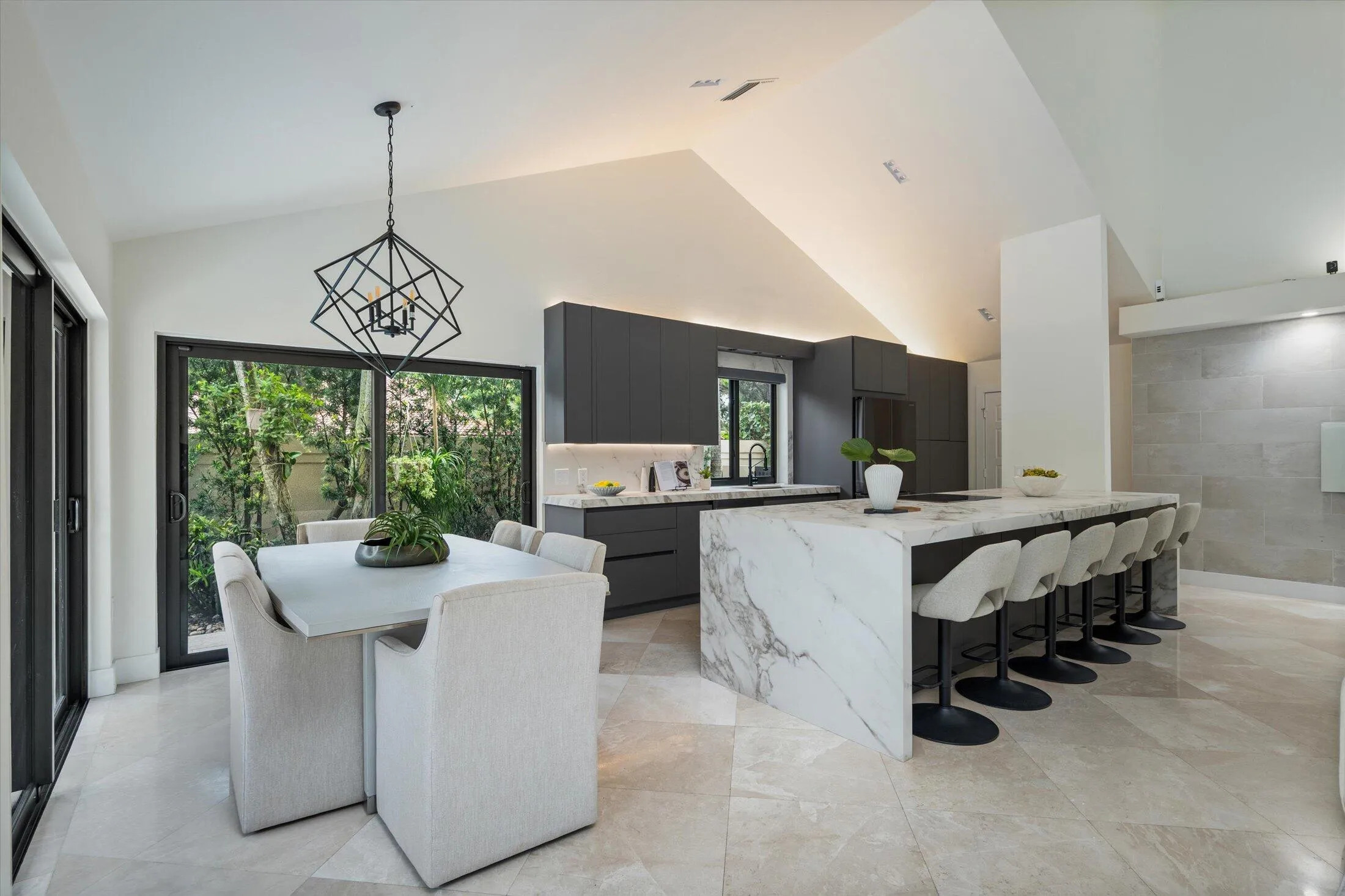 Vaulted ceilings with recessed lighting, large format floor tiling, a waterfall-edge island, dark flat-panel cabinetry, and sliding glass doors