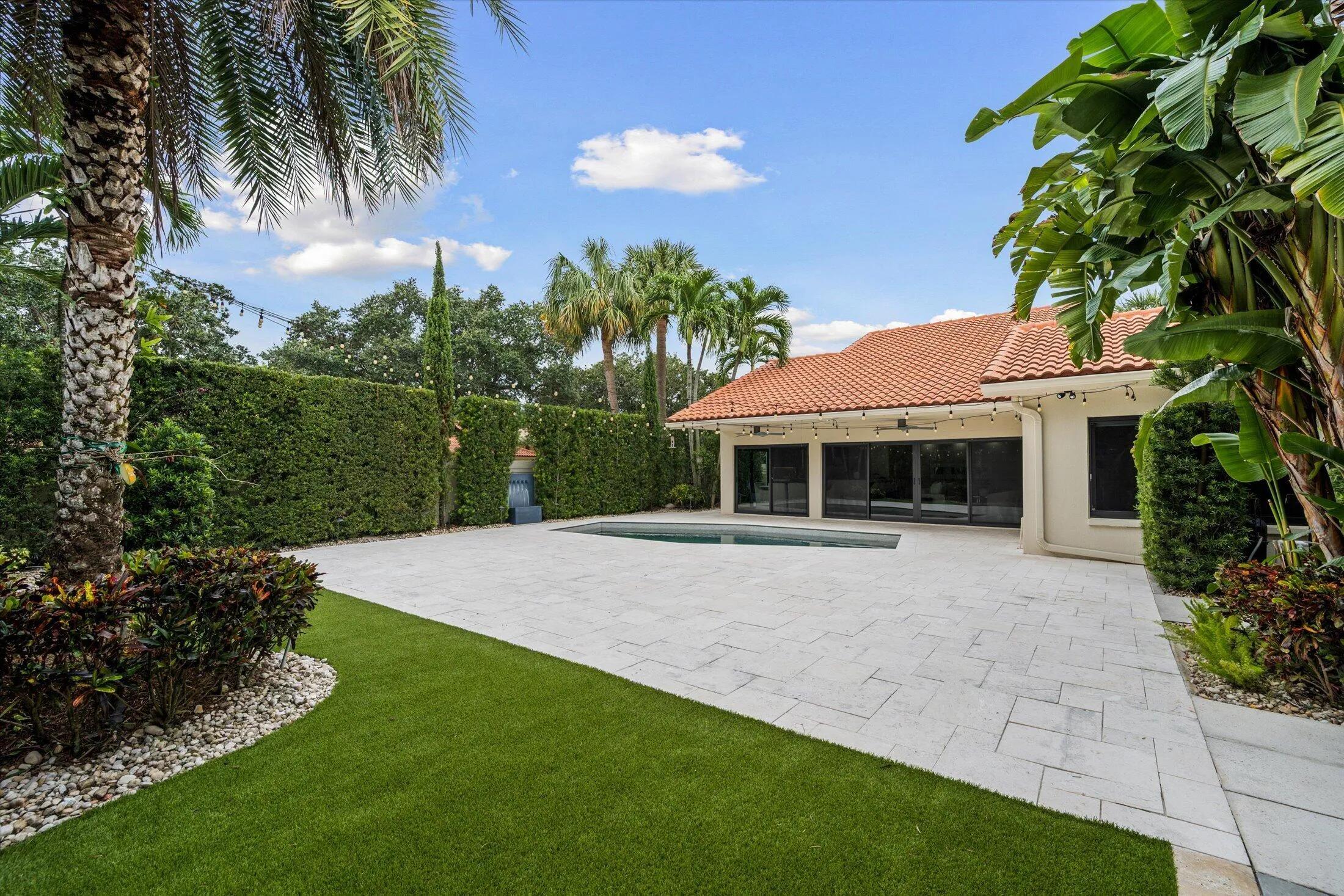 Expansive paver patio with a rectangular pool, artificial turf lawn, and mature tropical landscaping
