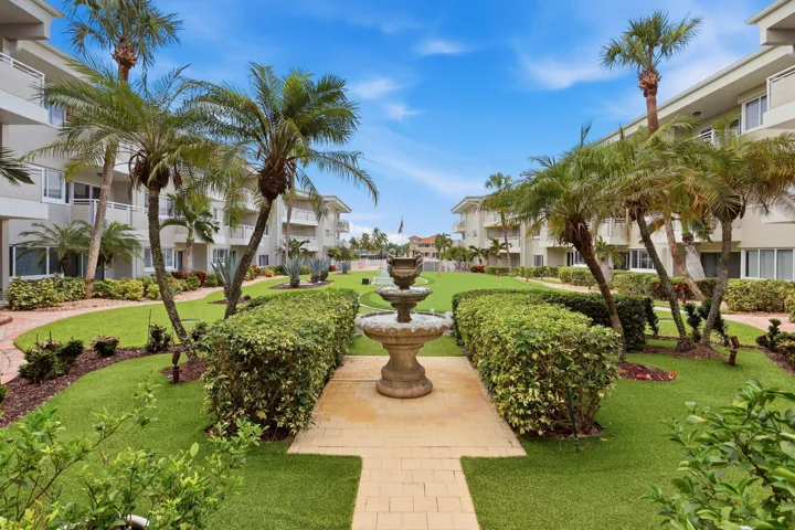 Landscaped courtyard featuring a tiered fountain, mature palm trees, manicured hedges, and paver pathways