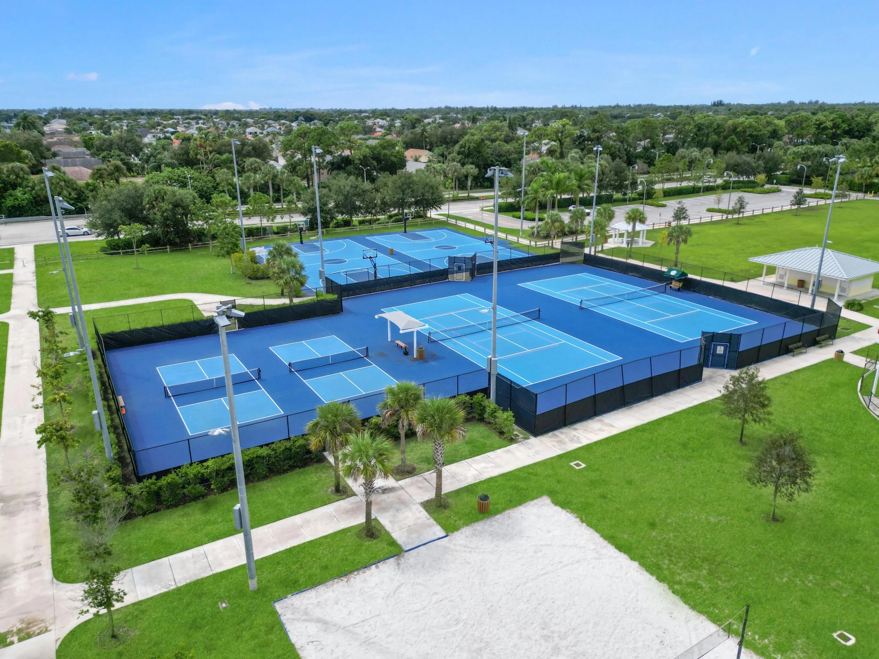 Multiple blue hard courts with perimeter fencing, surrounded by manicured green spaces and mature trees