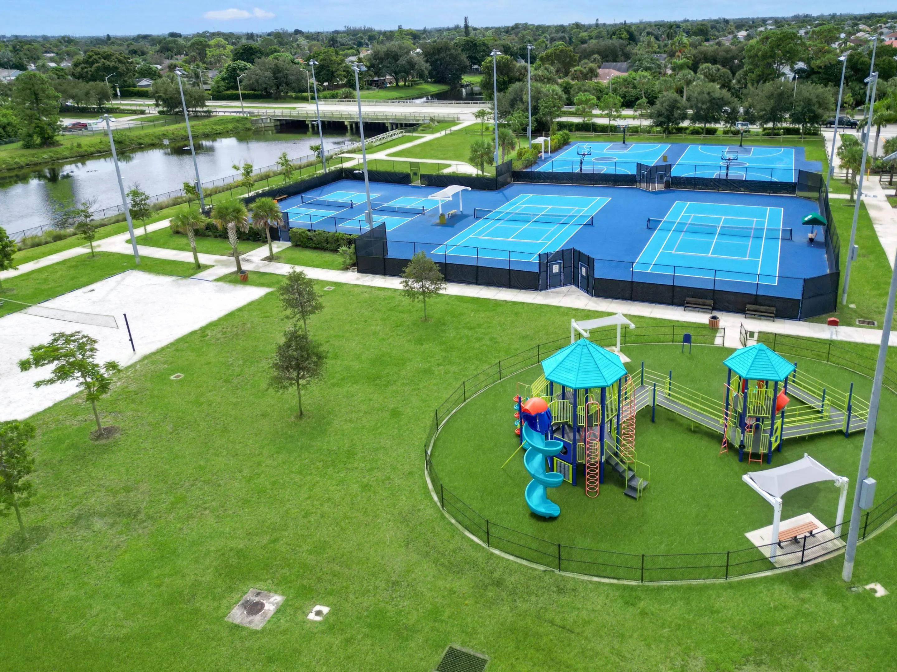 Recreational area featuring multiple blue-surfaced tennis courts, blue-surfaced basketball courts, a sand volleyball court, and a fenced playground with artificial turf