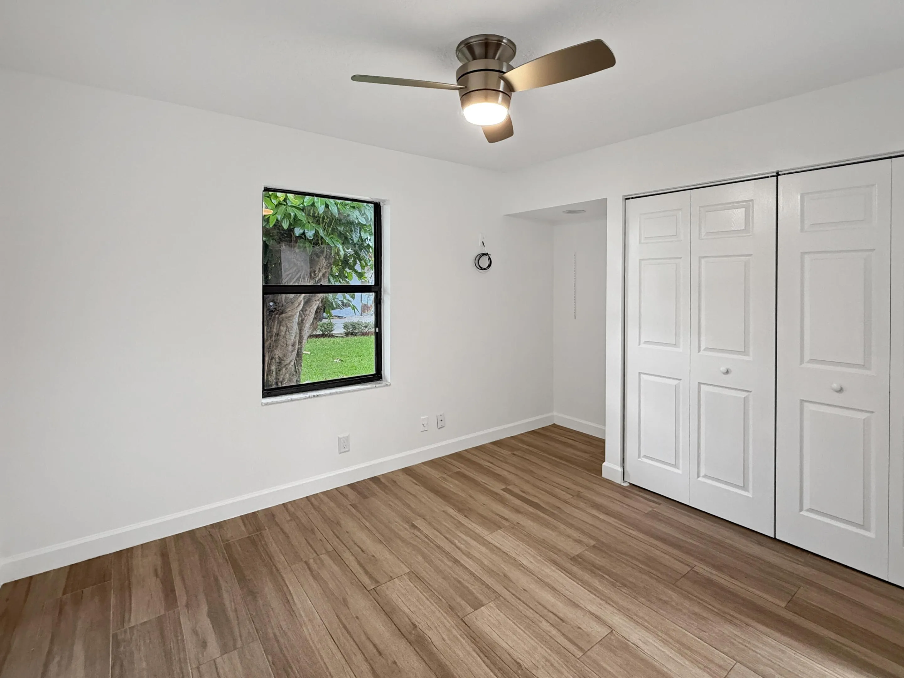 Spacious bedroom showcasing warm wood-finish flooring, white six-panel closet doors with multiple storage sections, ceiling-mounted fan with integrated lighting, and abundant natural light from a large window framing outdoor scenery