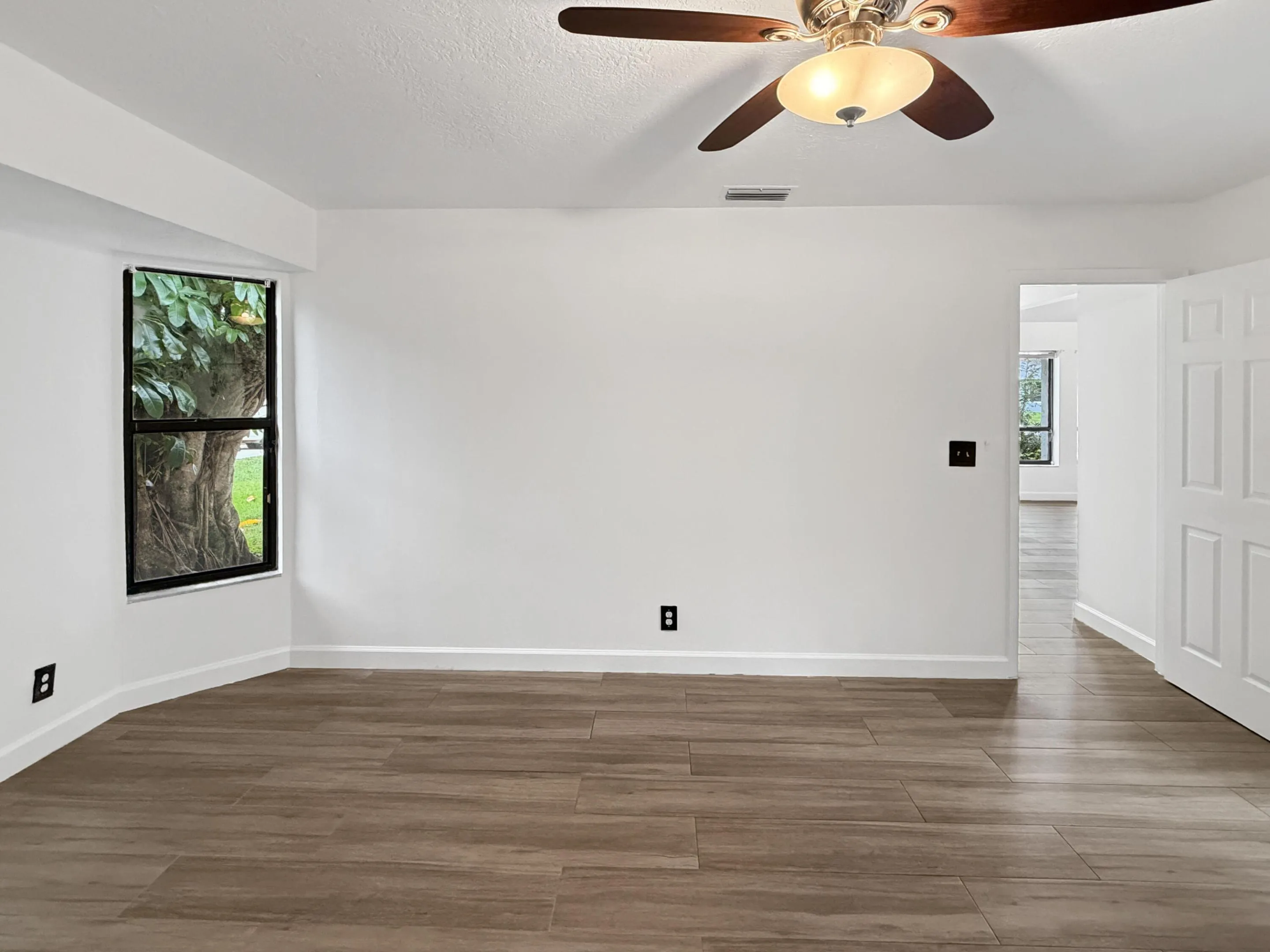 Wood-finish tile flooring, white painted walls, ceiling fan with integrated lighting, and a single window with exterior scenery