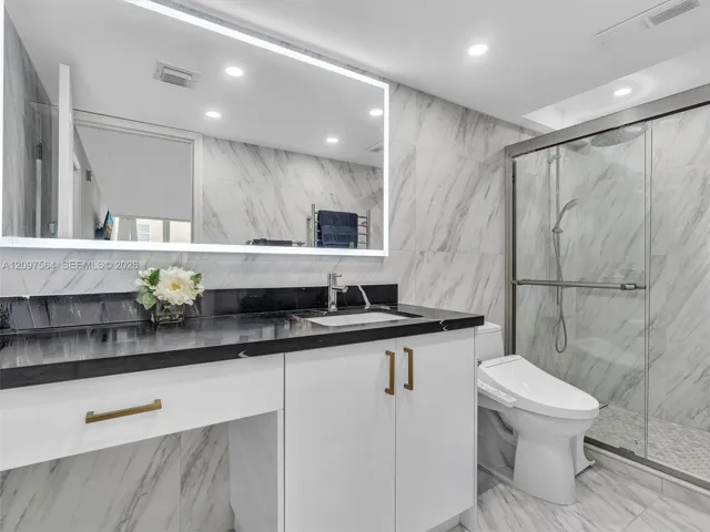 Stone-topped vanity featuring white cabinetry, metallic hardware, an integrated rectangular sink, and a large LED-backlit mirror