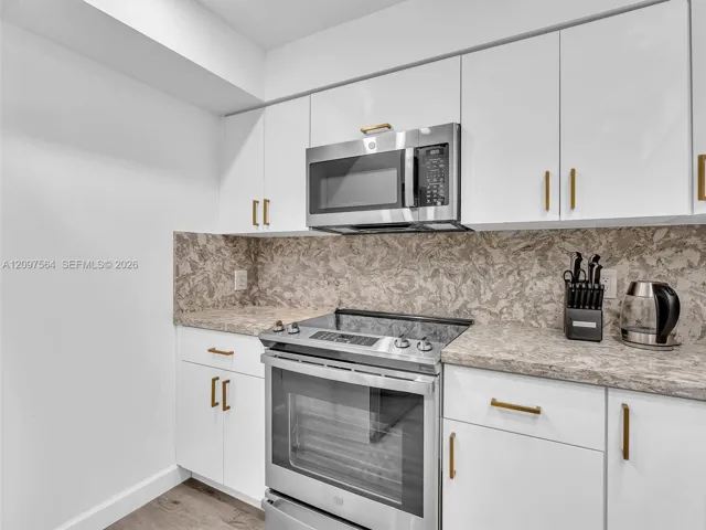 Kitchen featuring white flat-panel cabinetry, brushed gold hardware, stone-finish countertops, coordinating full-height backsplash, and stainless steel appliances