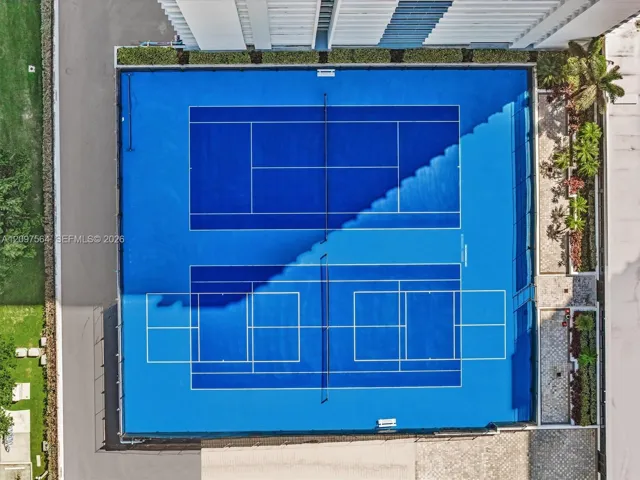 Aerial view of dual blue-surfaced courts featuring perimeter fencing, integrated net systems, and stone-paver walkway bordering native landscaping